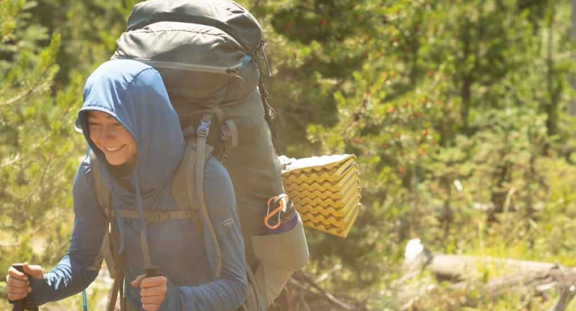 a student carrying a backpack smiles as they hike among trees on an outward bound course 